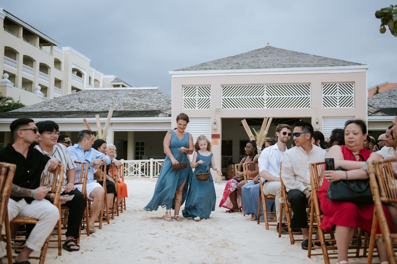 Wedding guests seated on wooden chairs during a beachside ceremony with a white gazebo in the background.