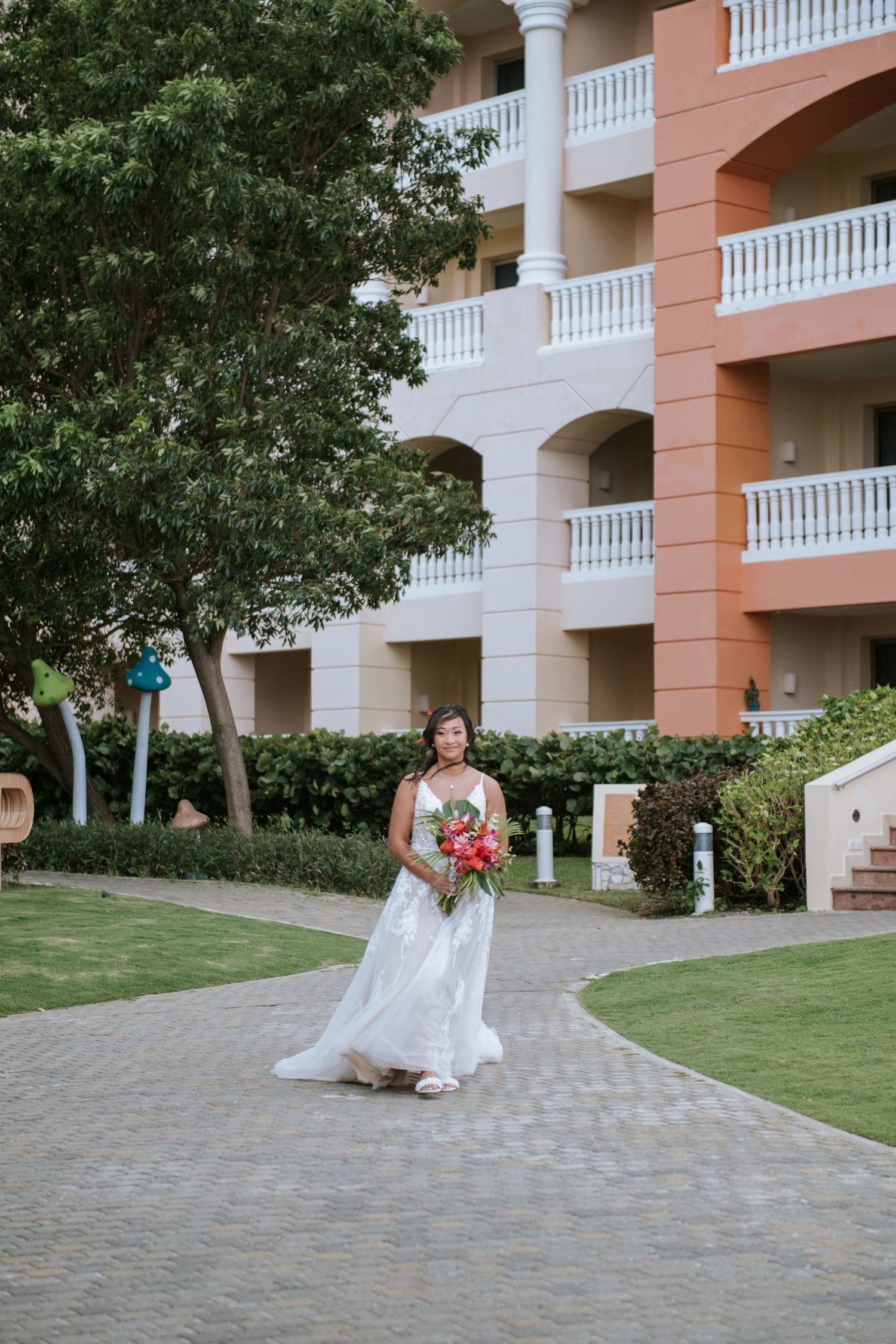 A person in a white wedding dress holding a red bouquet walks along a path near a coastal resort building.