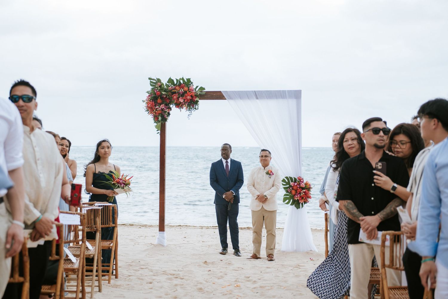 Beachfront wedding ceremony with red floral arch and wooden chairs facing the ocean.