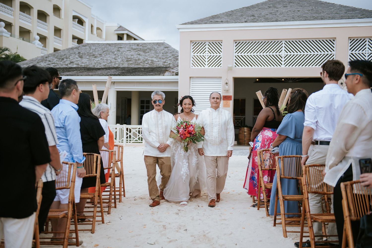 A wedding ceremony procession on a white sandy beach path with wooden chairs and guests.