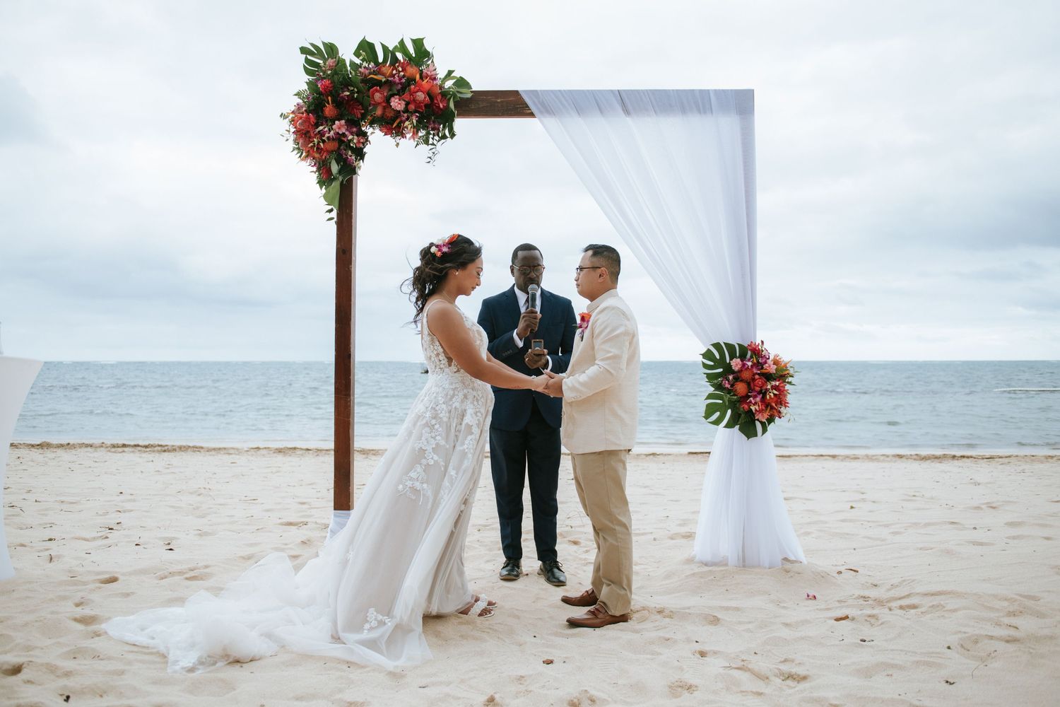 A wedding ceremony under a wooden arch with red flowers against an ocean backdrop.