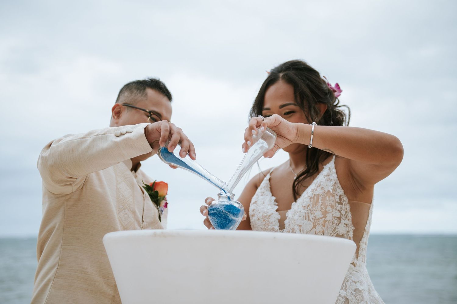 A couple performs a sand ceremony ritual with blue sand at their beach wedding.