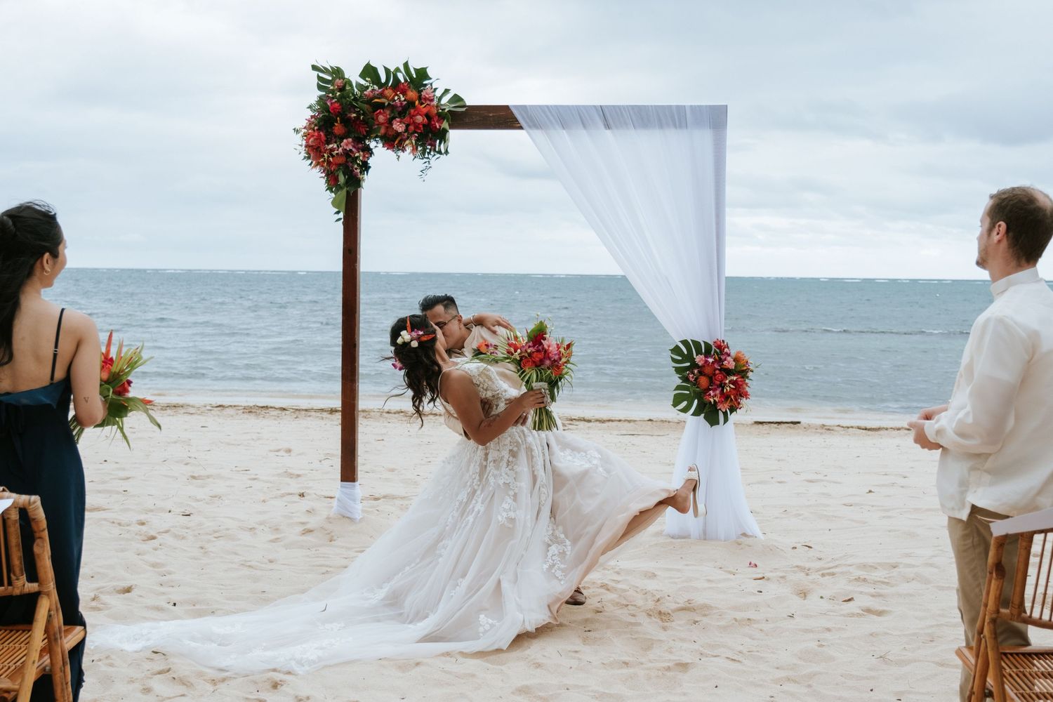 A romantic beach wedding ceremony setup with a white fabric arch decorated with tropical flowers against an ocean backdrop.