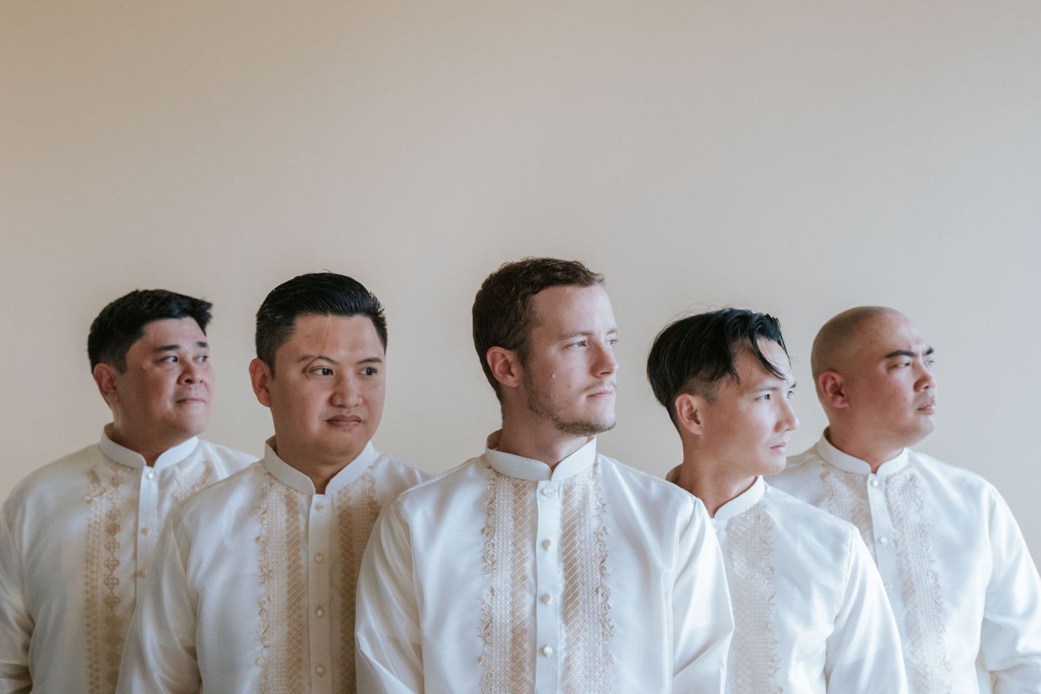 Five men in white dress shirts standing in line against neutral wall.
