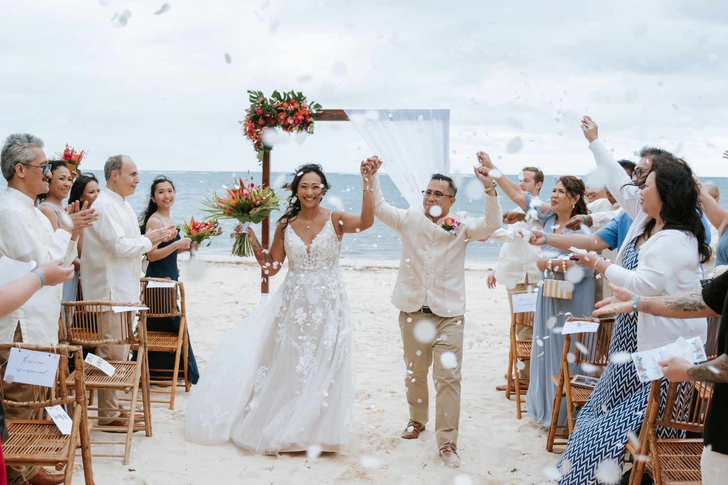 Wedding celebration on a beach with guests cheering as newlyweds walk down the aisle.