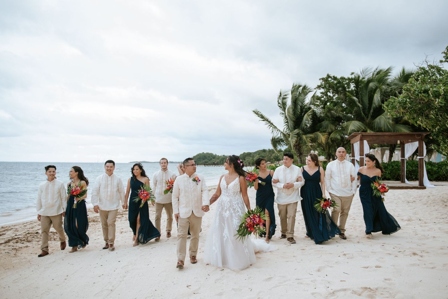 Wedding party poses for photos on a beach with palm trees and ocean in the background.