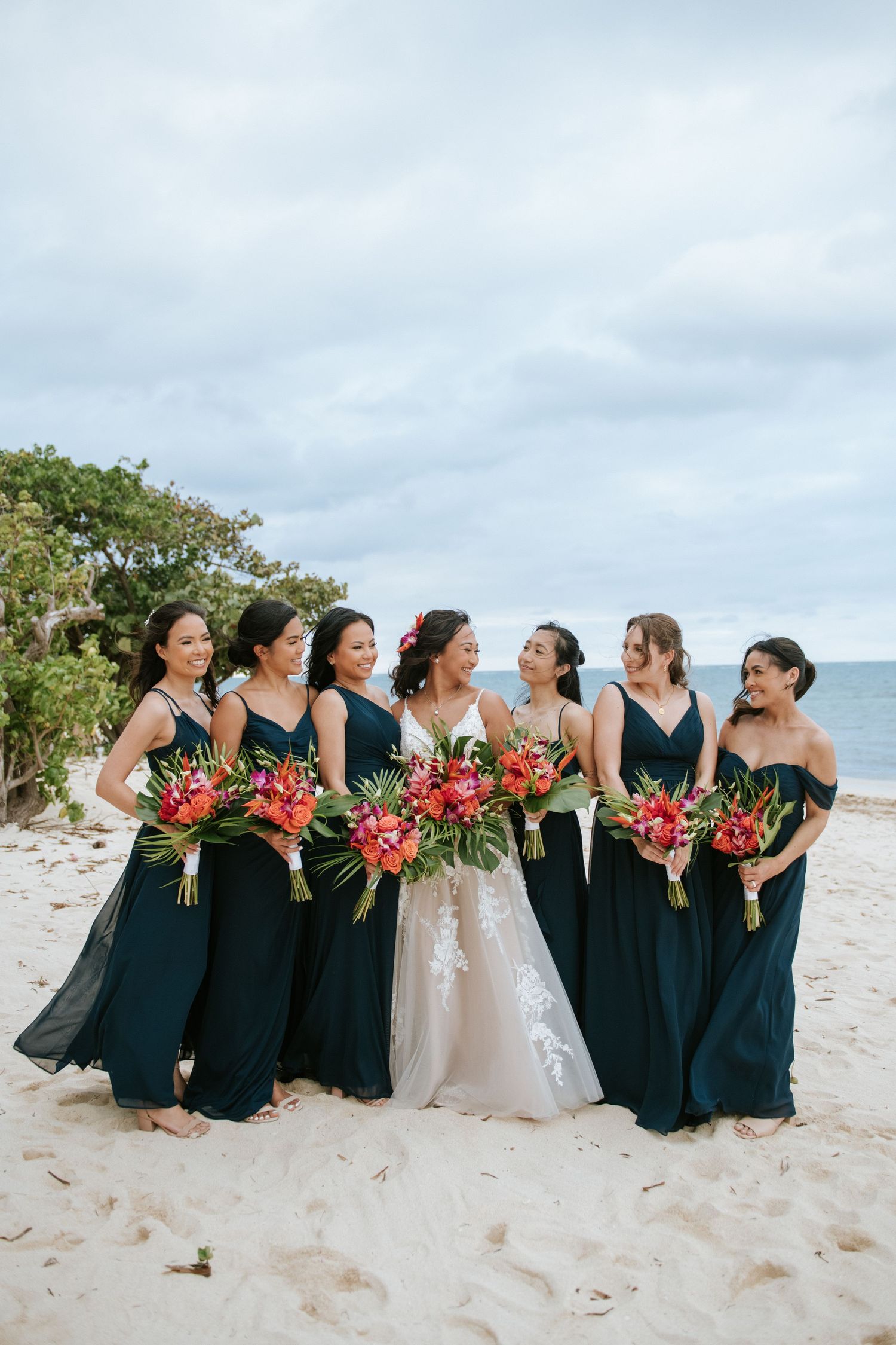 Bridal party in elegant navy blue dresses holding vibrant tropical bouquets pose together on a sandy beach.