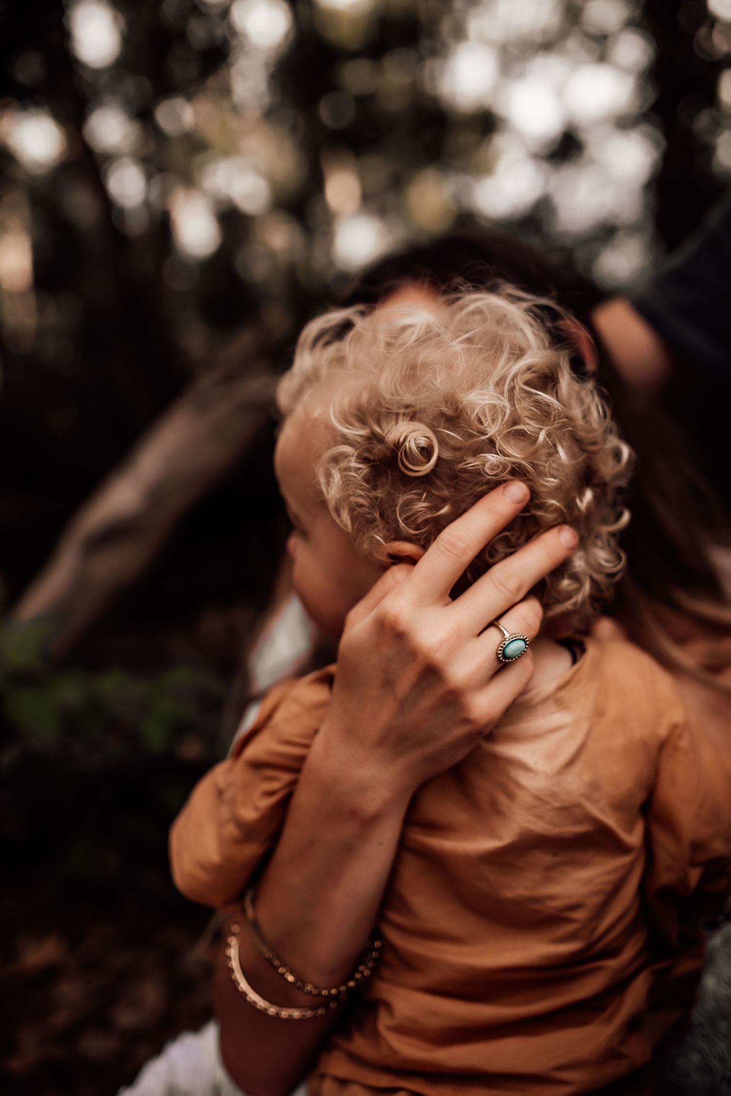 A hand with a ring gently touches blonde curly hair in a moody outdoor setting.