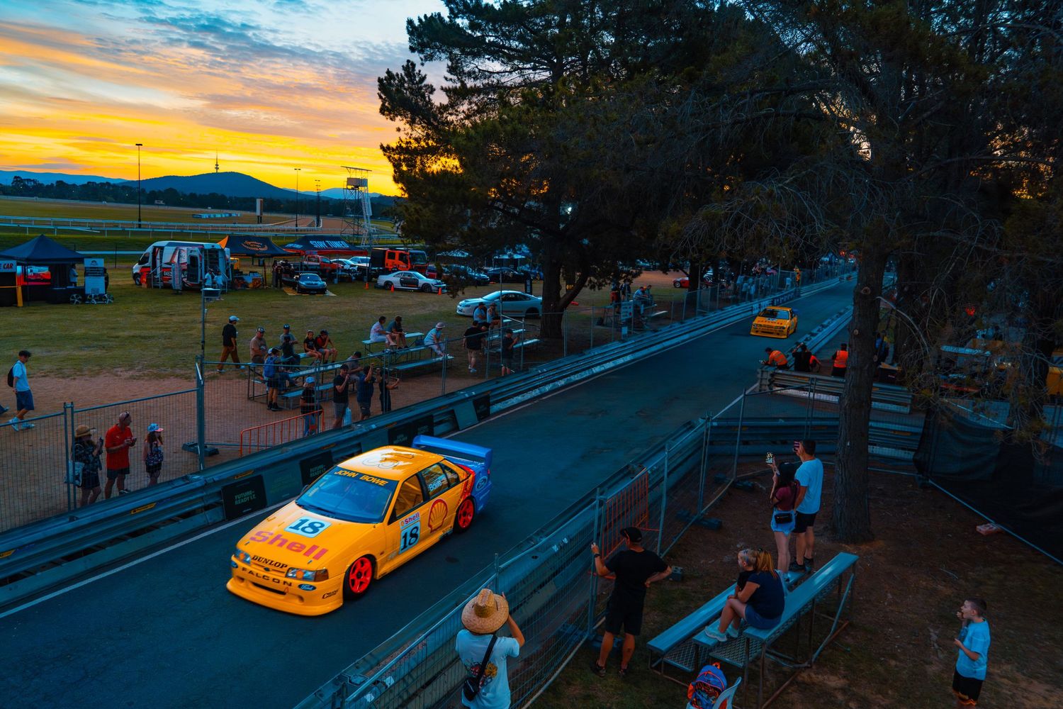 Yellow race car on track during sunset with spectators watching from hillside.