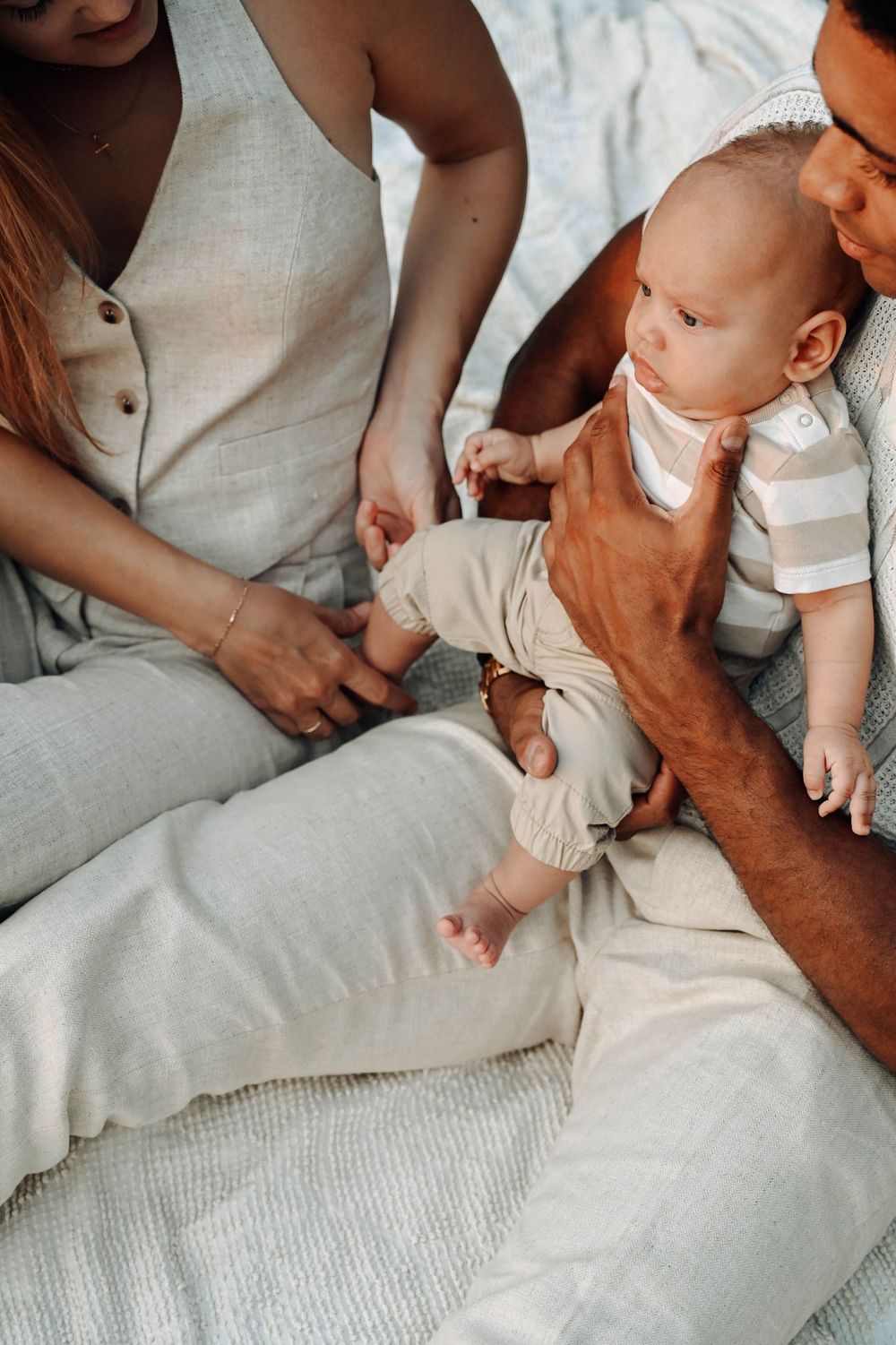 Baby in light colored clothing lies between parents on white bedding during intimate family moment.