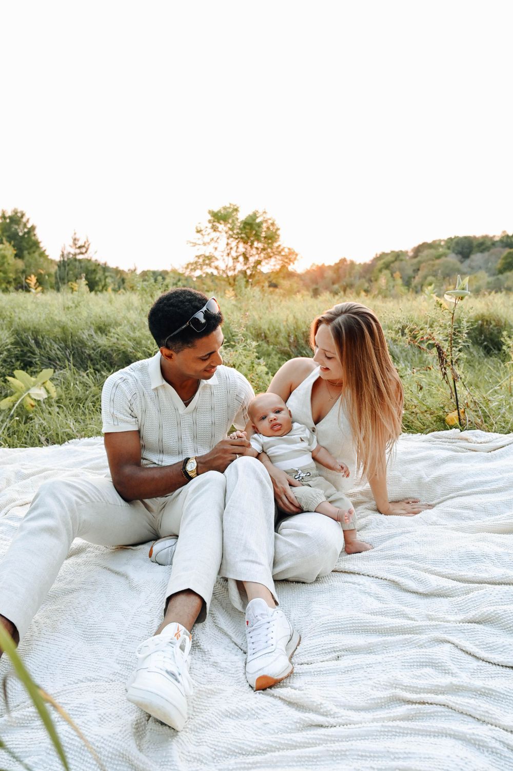 A couple sits together on a white blanket in a sunlit meadow during golden hour, enjoying a peaceful outdoor moment.