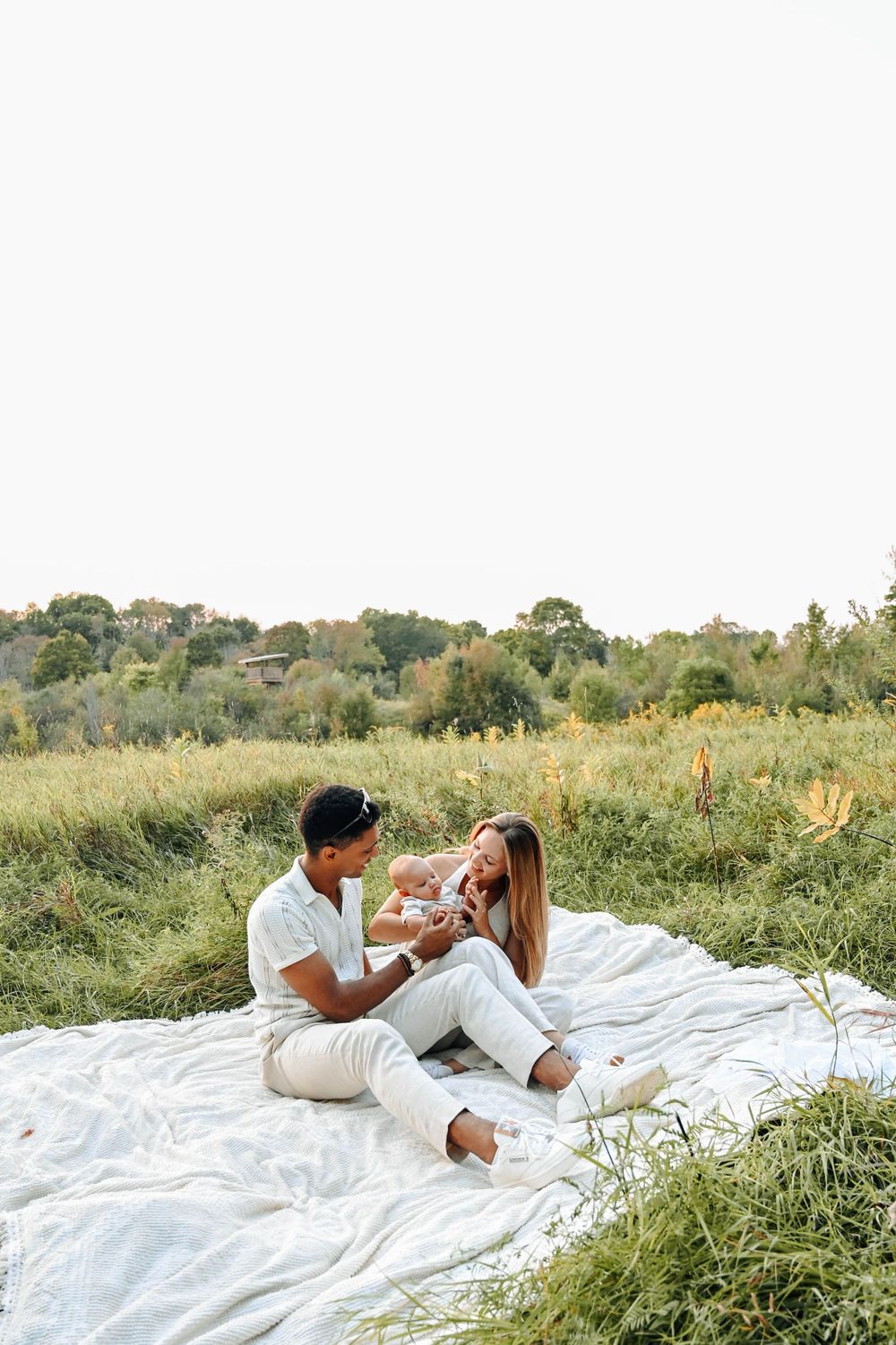A couple sits on a white blanket in a grassy meadow during golden hour enjoying a picturesque moment together.