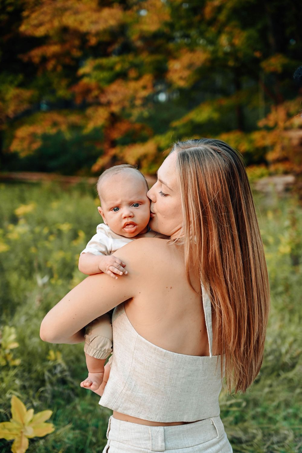 A tender moment captured outdoors with autumn foliage as a baby is held and kissed in a natural setting.