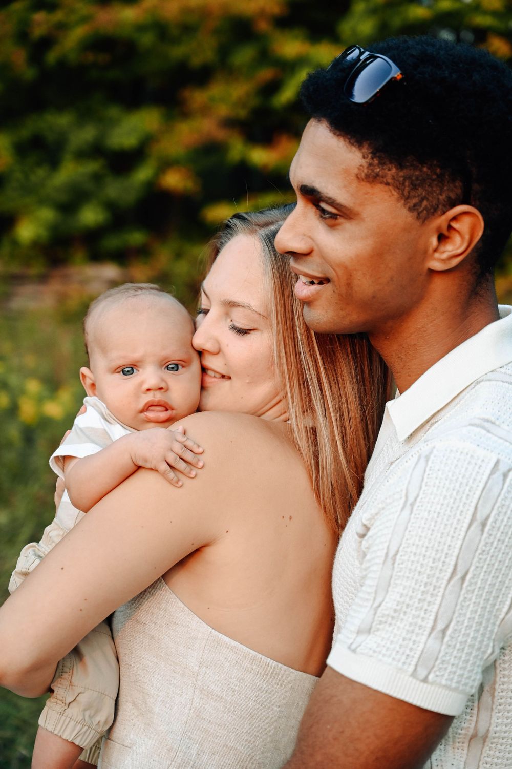 A loving family embraces outdoors, with a small baby and parents in casual white clothing against a green background.