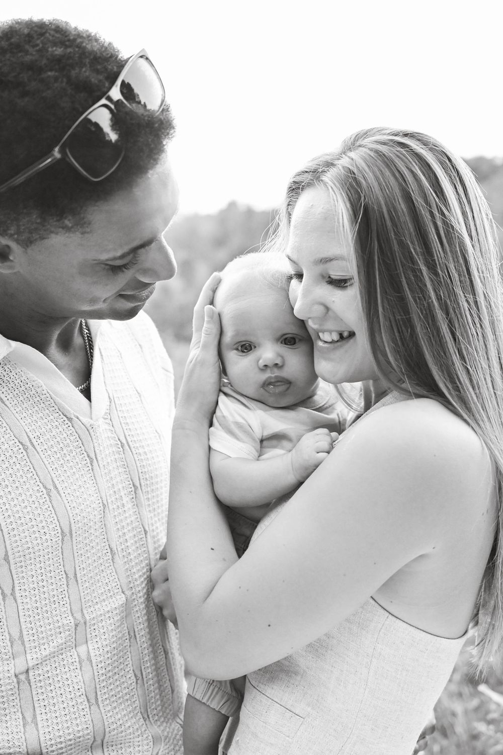 A loving couple shares a tender moment with their baby outdoors in a black and white portrait.