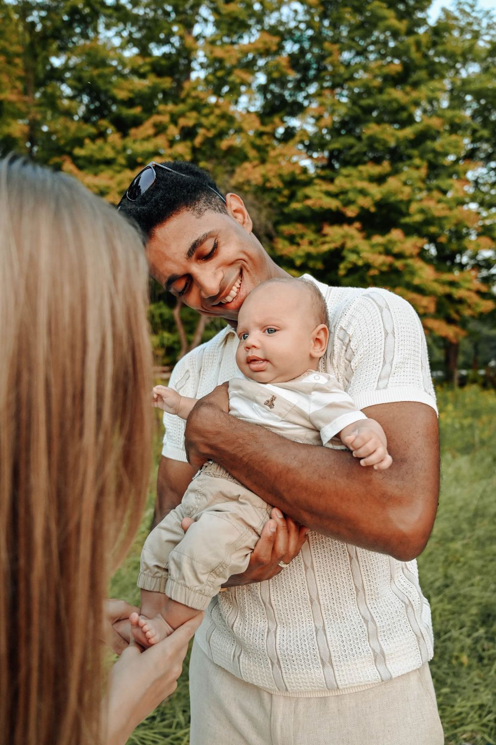 A baby in khaki pants and white shirt is held lovingly outdoors against an autumn backdrop of trees.