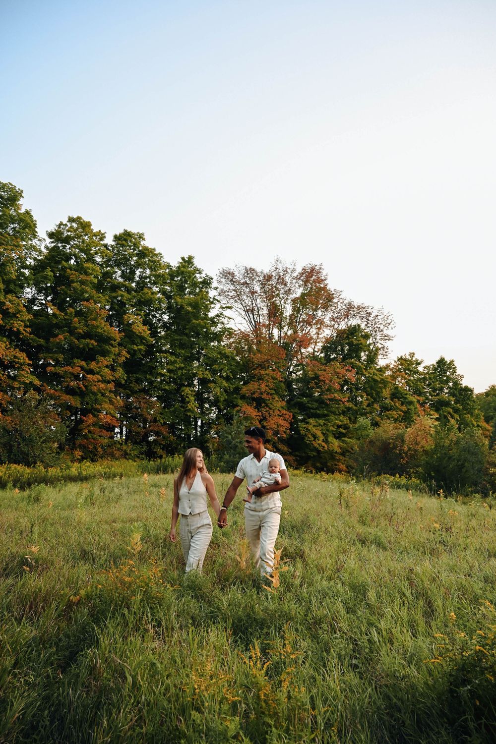 A couple walking through a sunlit meadow surrounded by autumn trees.