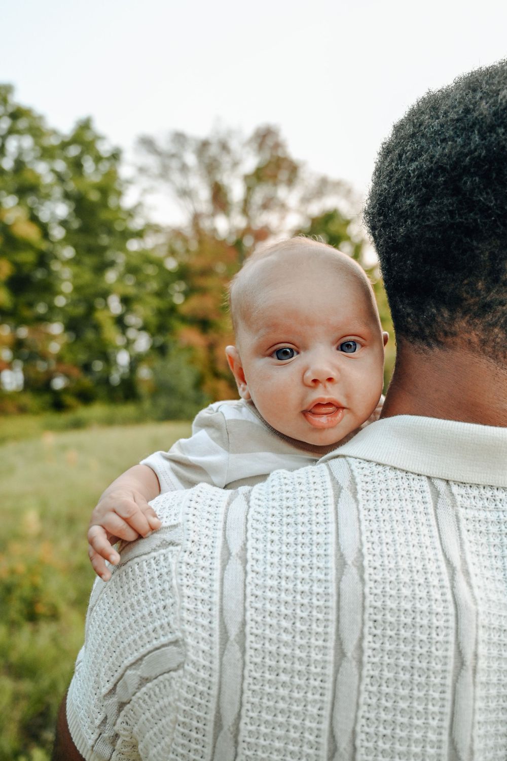 A baby peers over someone's shoulder wearing a white sweater in an outdoor setting with trees in the background.