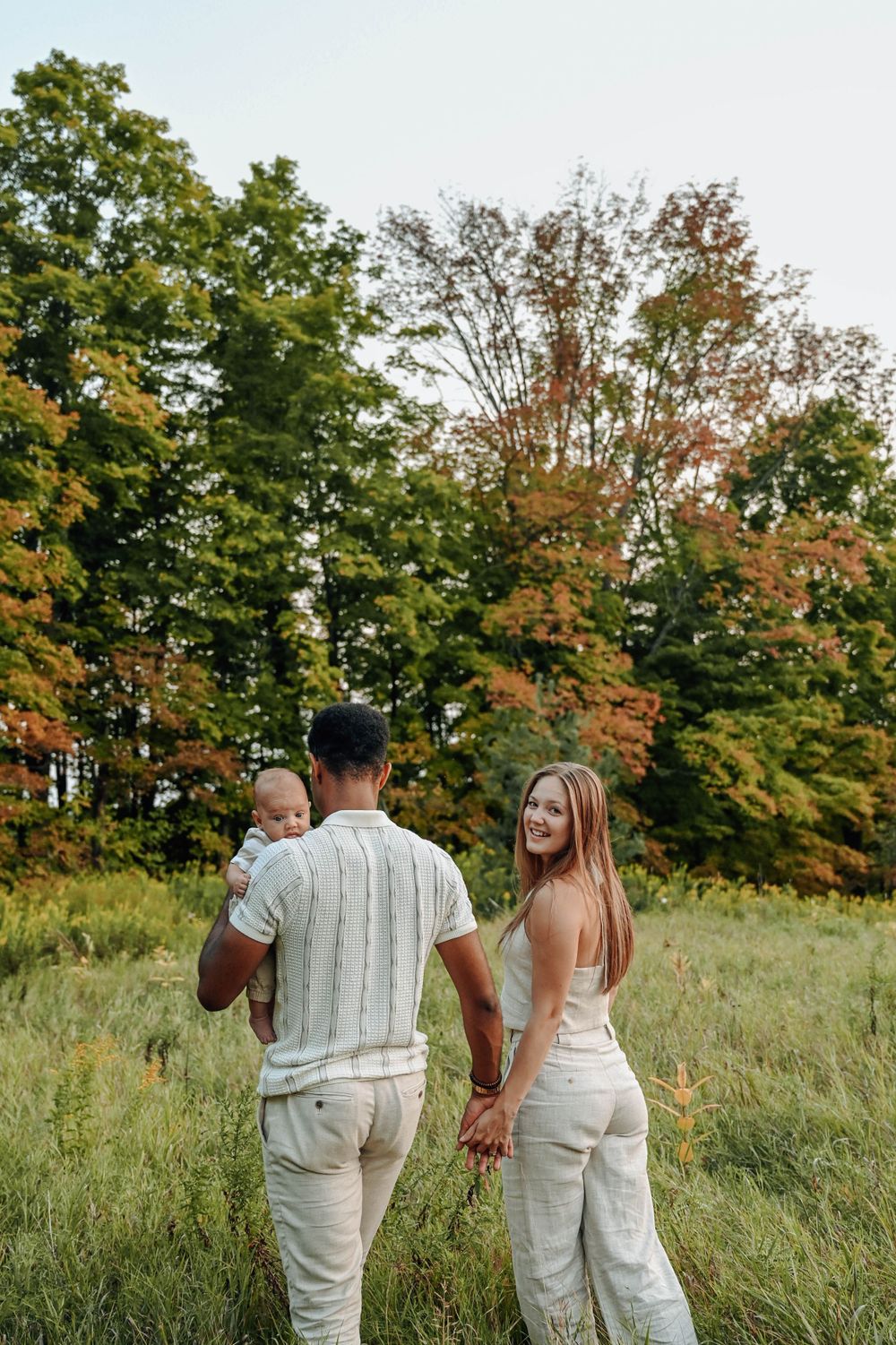 A couple walking through a field with autumn trees showing colorful foliage in the background.