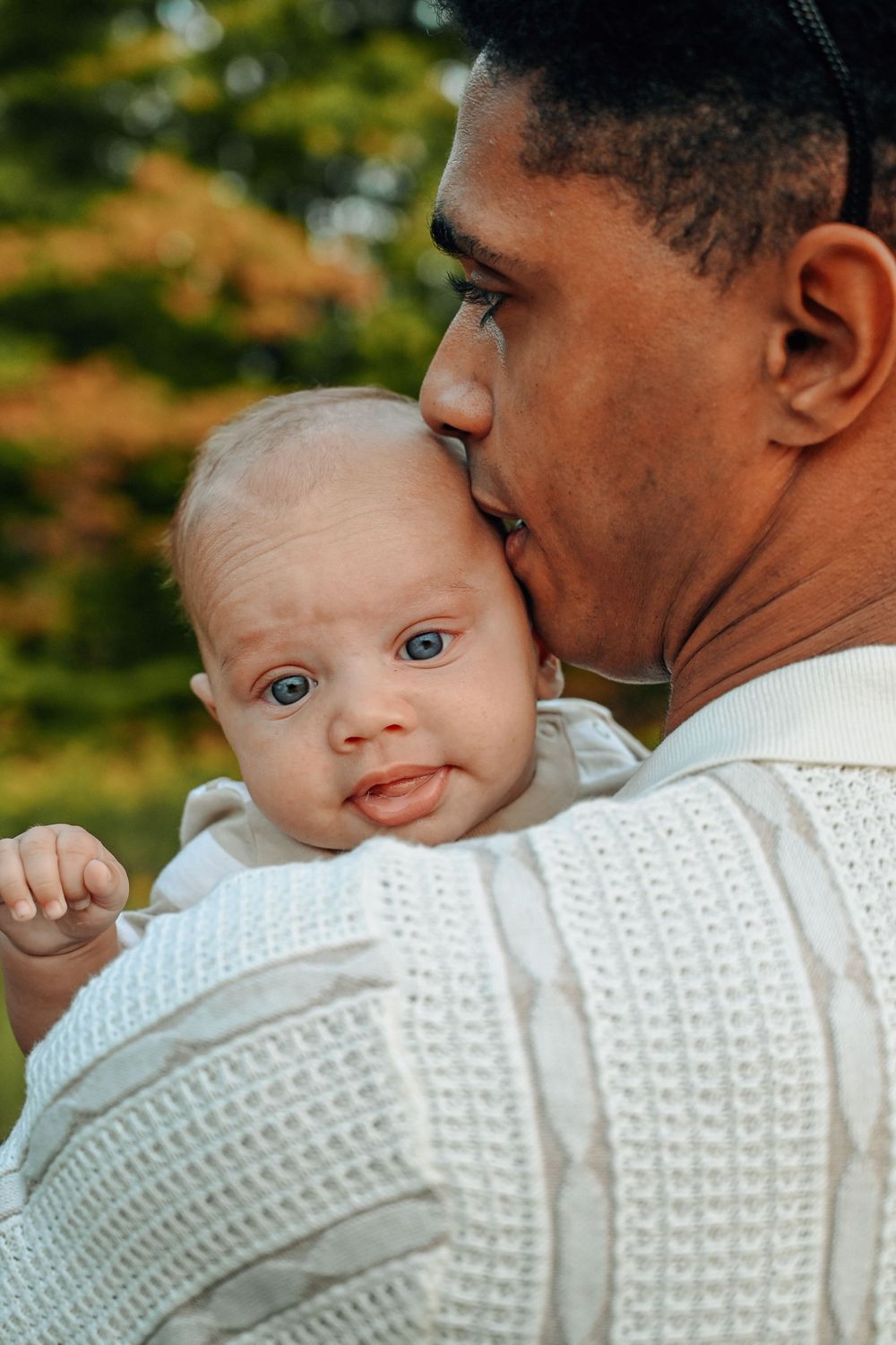 A tender moment between parent and baby in white knit sweaters against an autumn background.