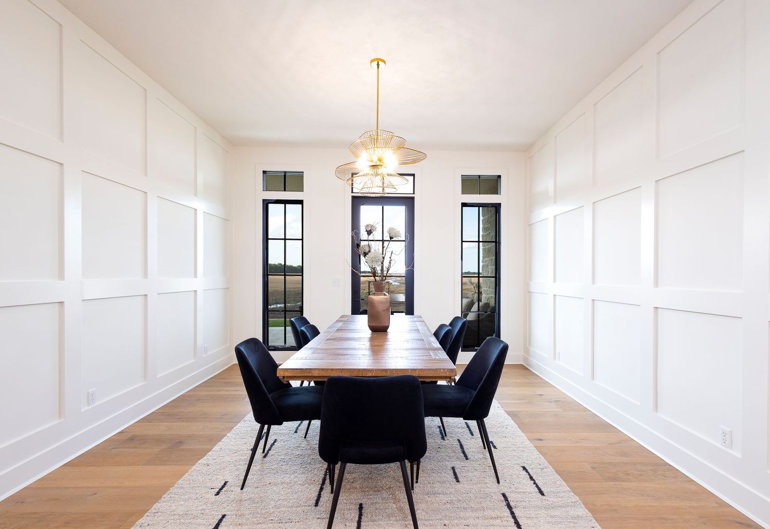 Modern white dining room with black velvet chairs, hardwood floors, and a brass chandelier illuminating the space.