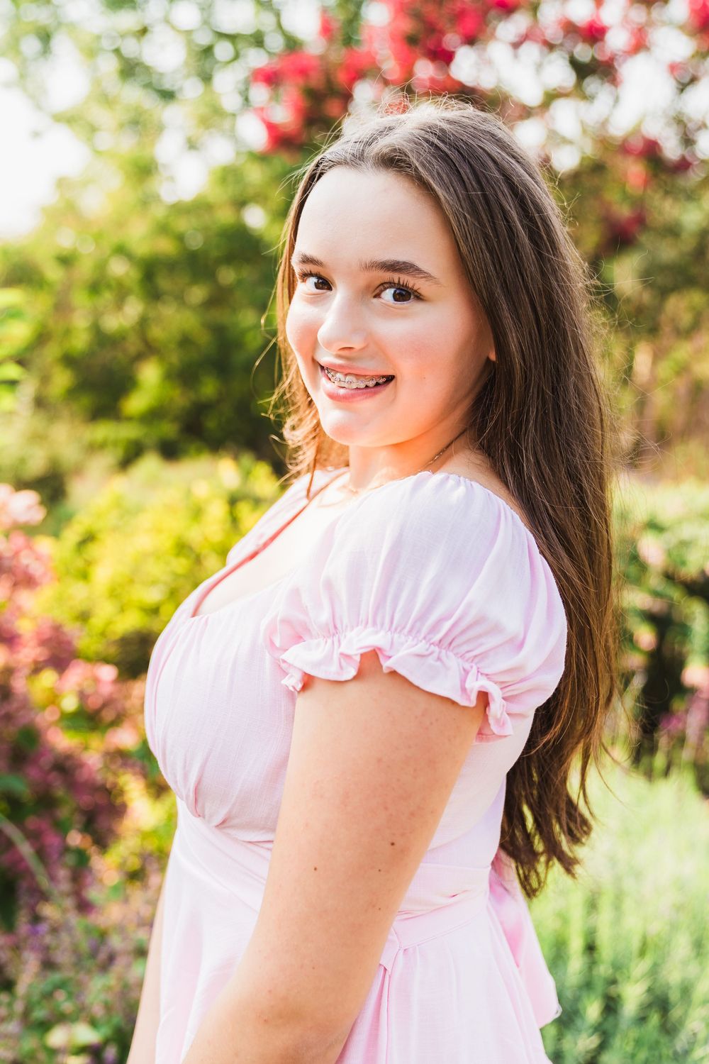 Smiling teenage girl with braces in a pink dress posing outdoors in a garden with colorful flowers.