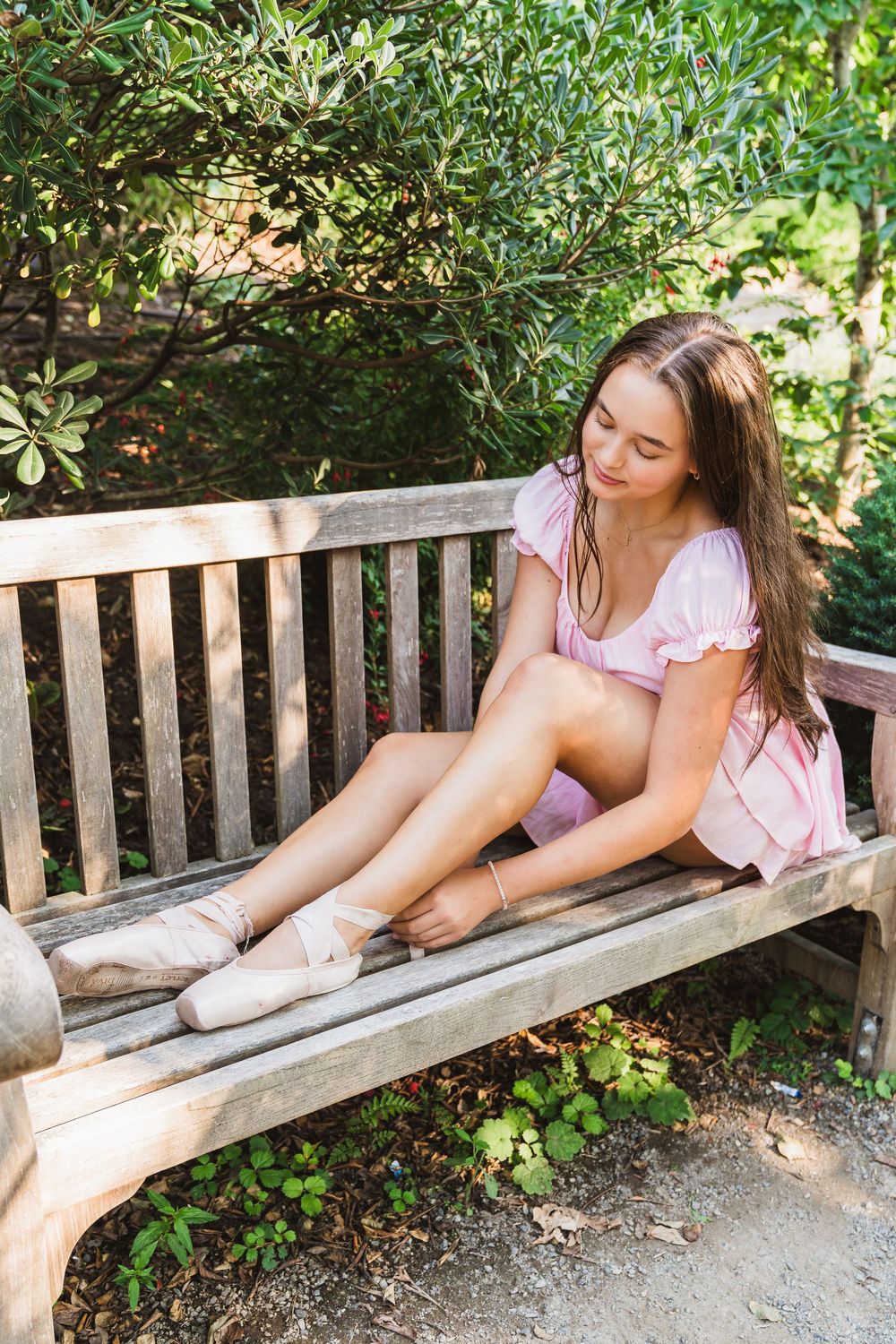 Young girl in pink dress sitting on wooden bench putting on ballet pointe shoes outdoors.