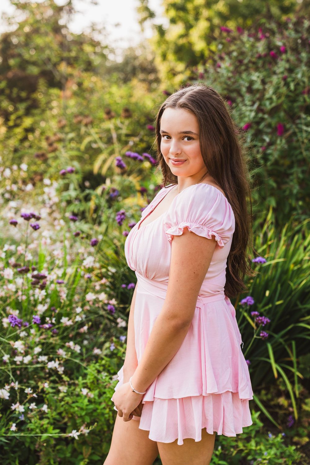Young woman in pink ruffle dress posing in a lush garden with purple and white wildflowers.