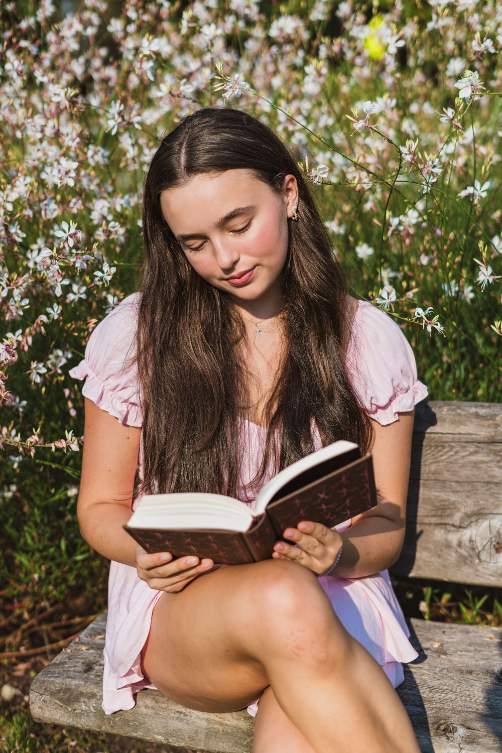 Young woman in pink dress reading a book on a wooden bench surrounded by blooming white flowers outdoors.