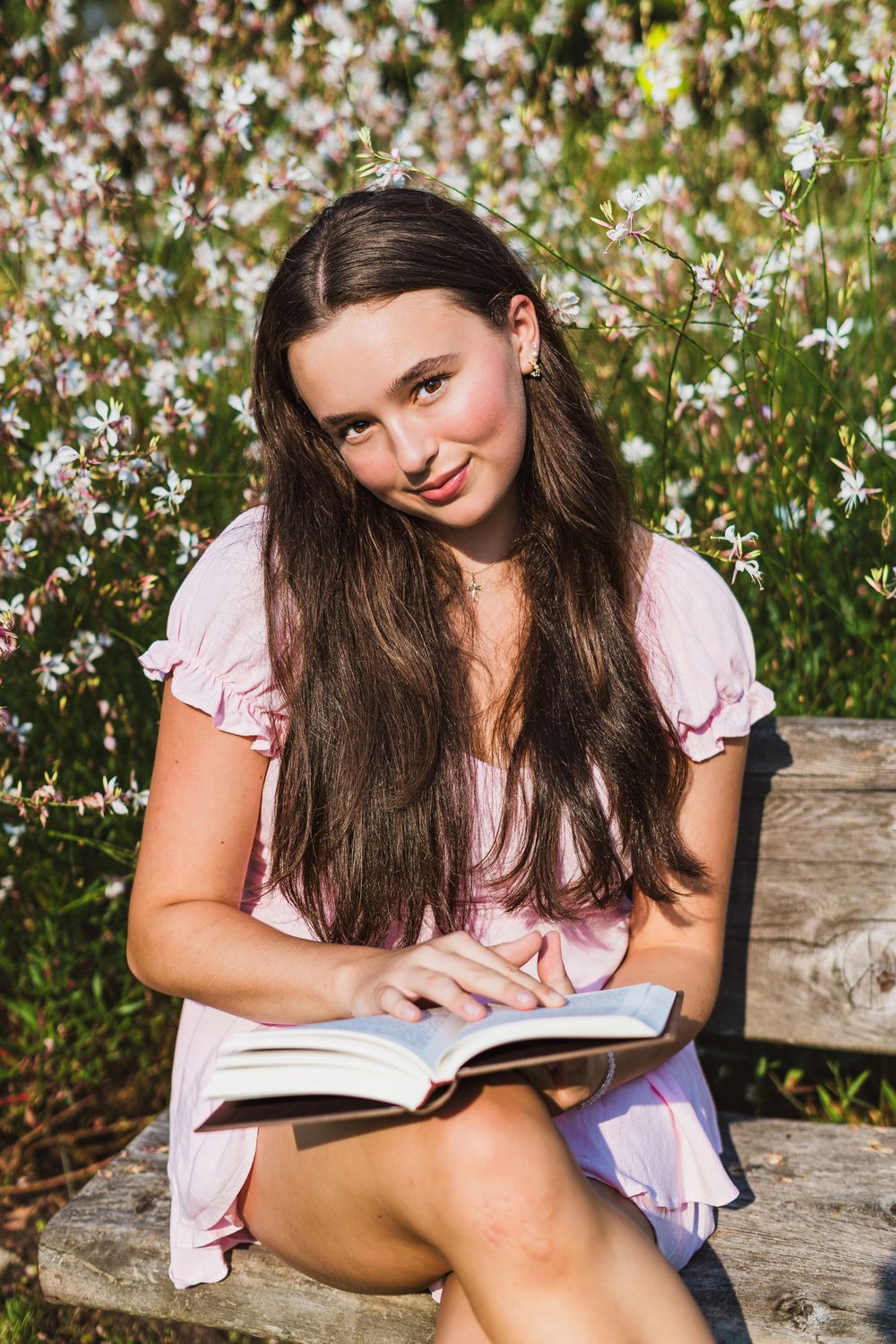 Teenage girl in pink dress reading book on bench surrounded by blooming white wildflowers.