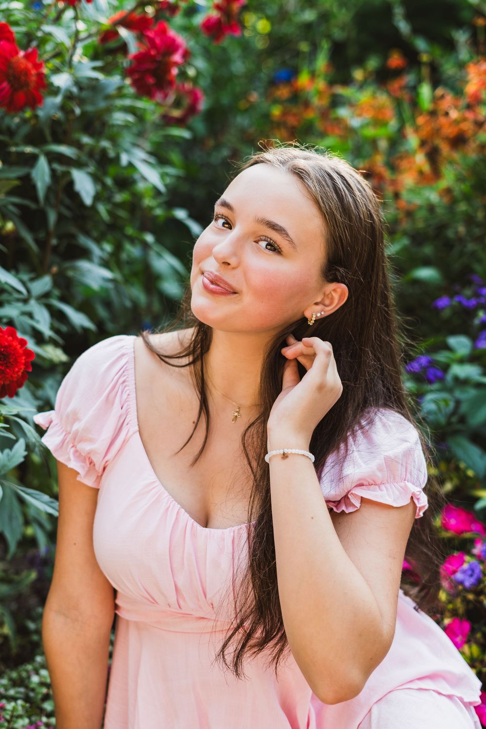 Young woman in pink dress posing outdoors among colorful garden flowers, smiling softly with hand near face.