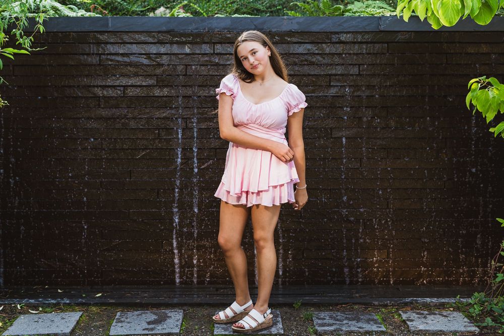 Young woman in pink mini dress and sandals posing in front of a dark wooden water feature wall.