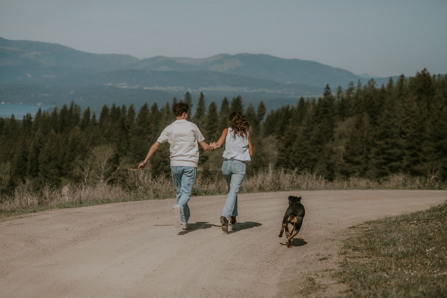 Couple holding hands running on a dirt road with a black dog, surrounded by pine trees and mountain views.