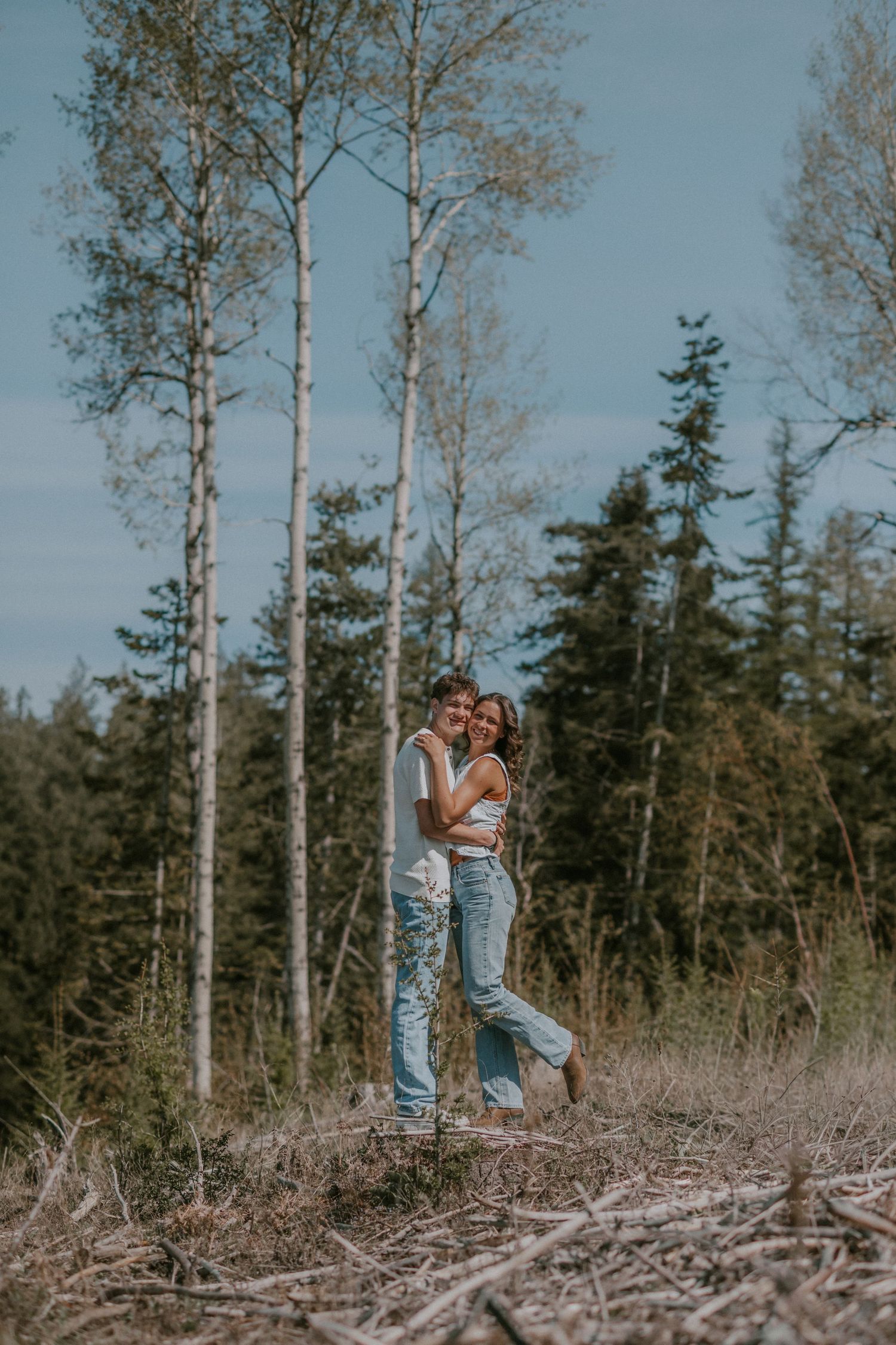Couple embracing and smiling in a forest clearing with tall birch and pine trees under a blue sky.