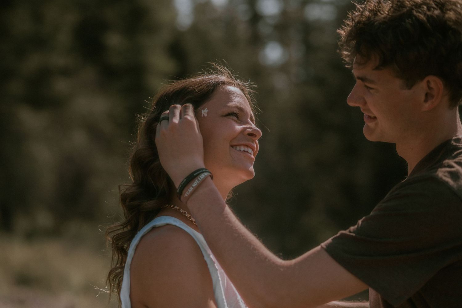 A young couple shares a tender moment outdoors, as a man gently brushes hair from a smiling woman's face.