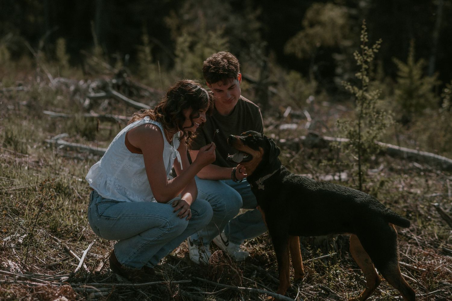 A young couple crouches in a forest petting a black and tan dog surrounded by fallen branches.