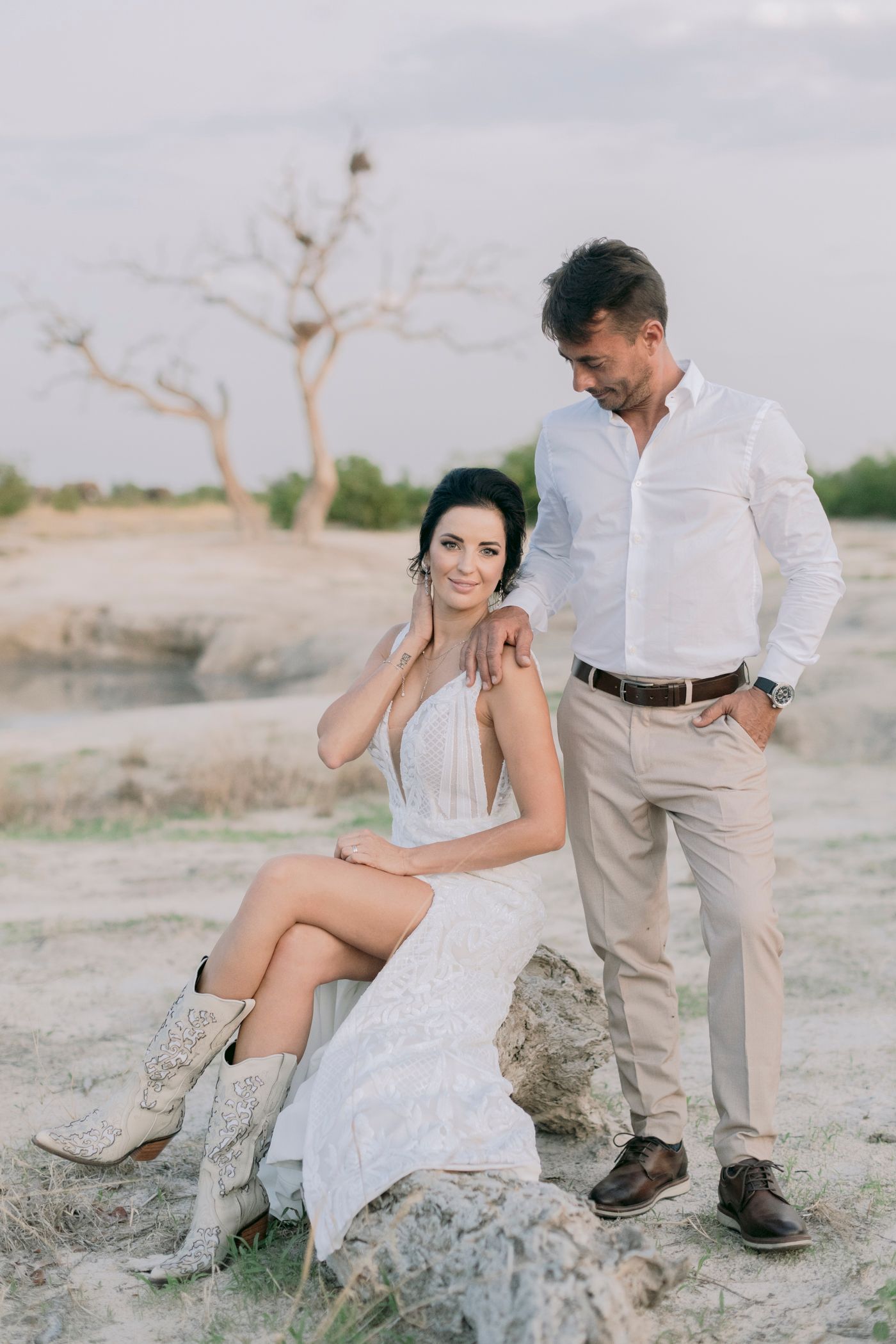 Couple posing in a rustic desert landscape, bride in white lace dress and cowboy boots with groom.