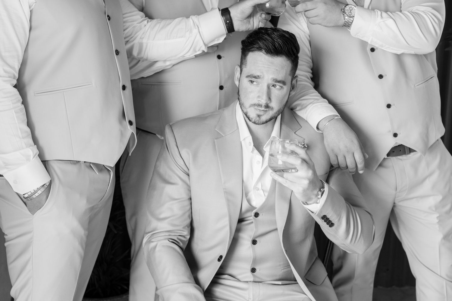 Groom sitting in light suit holding whiskey glass while groomsmen assist with preparations in black and white photo.