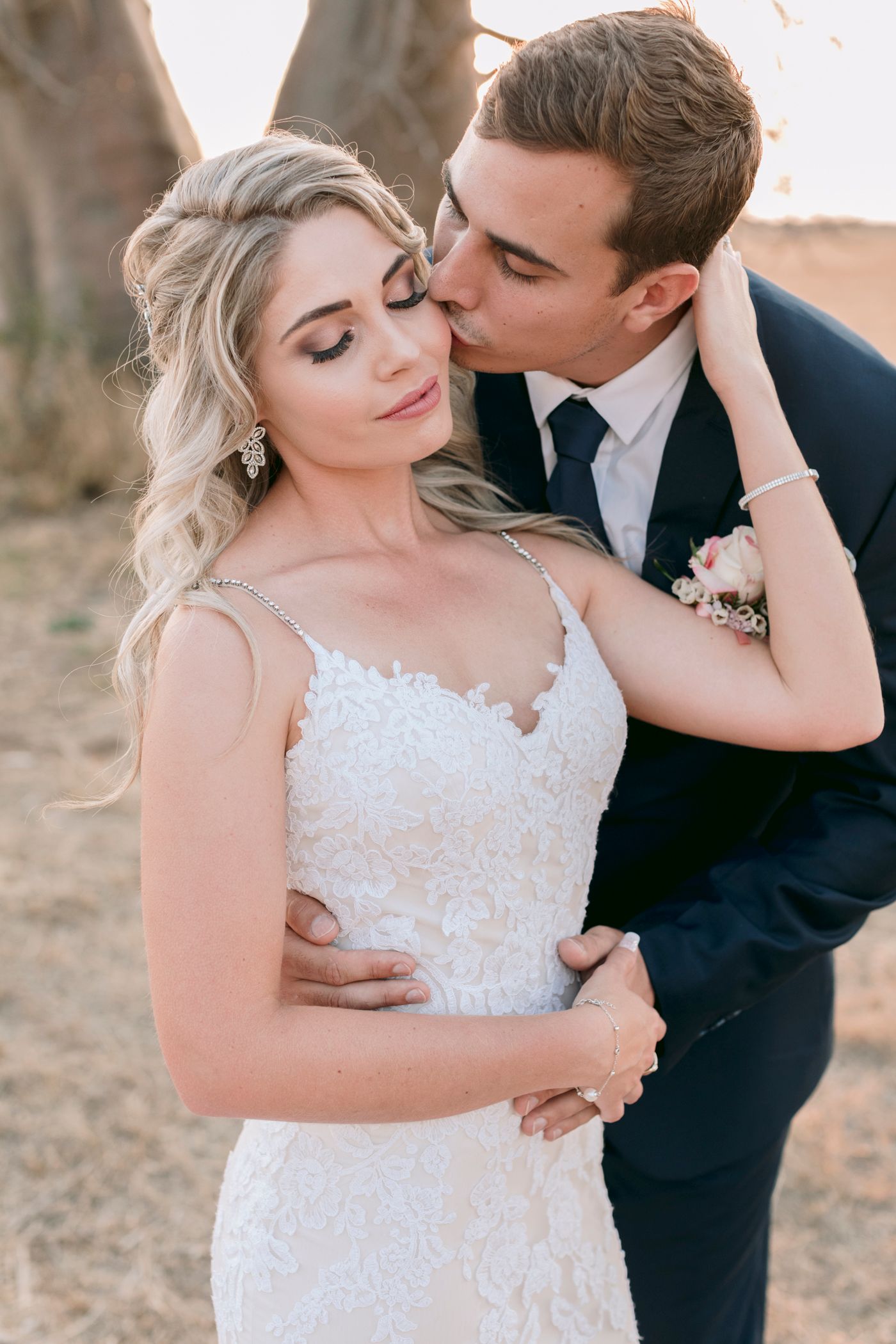 Groom in navy suit kissing bride on cheek as she smiles in lace wedding dress at golden hour.