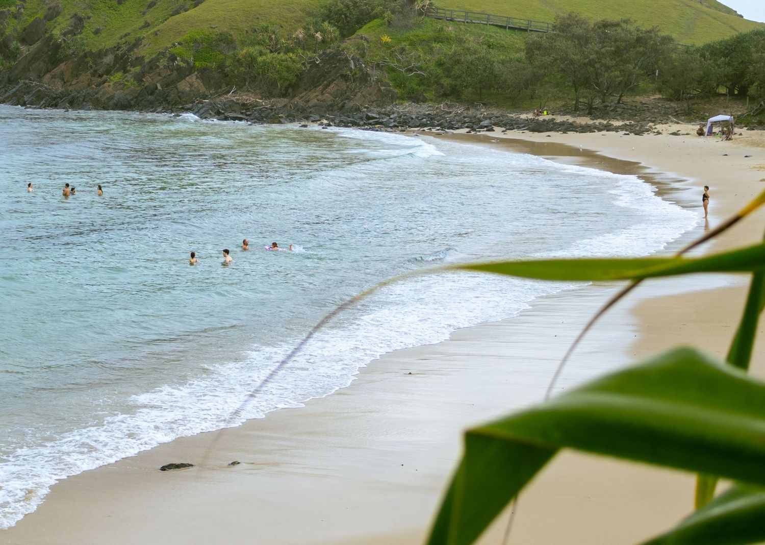 Swimmers enjoy calm turquoise waves at a scenic curved sandy beach framed by lush green tropical foliage.