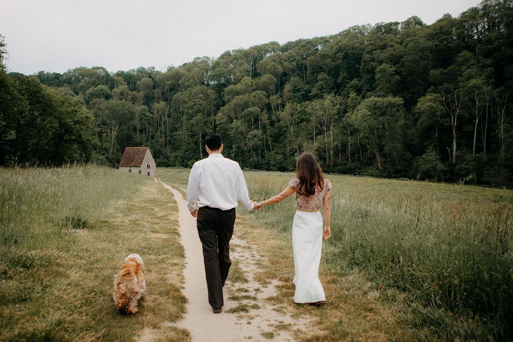 Bride and groom walking hand in hand with their golden dog along a meadow path.
