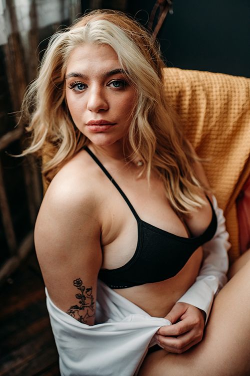 Blonde woman in black bralette and open white shirt with rose tattoo posing in chair.