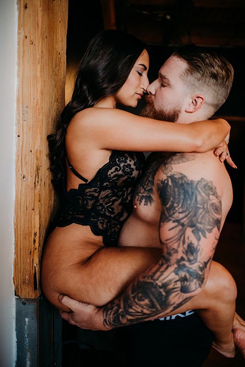 A tattooed man holds a woman in black lace lingerie against a wooden beam in an intimate romantic embrace.