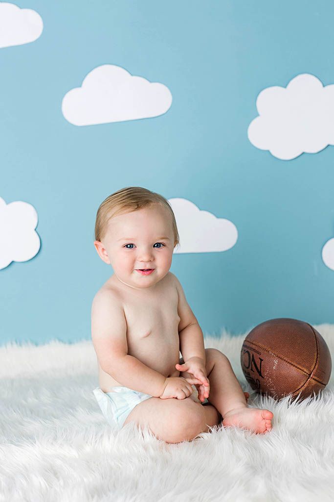 Smiling baby in diaper sits on white fur rug beside vintage football, blue backdrop with white clouds.