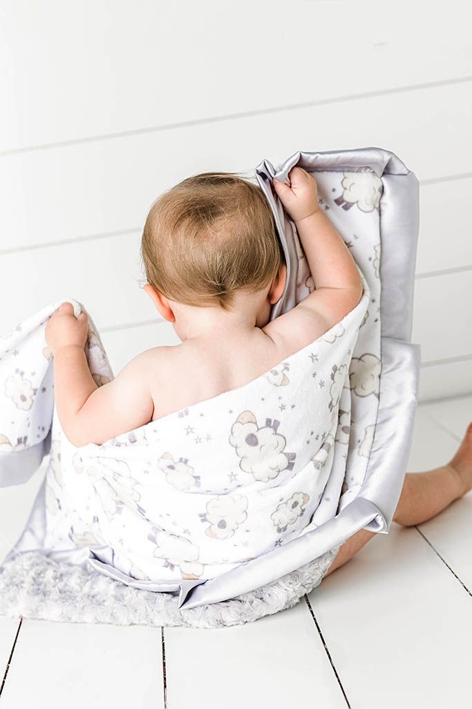 Baby wrapped in floral gray and white blanket sitting on white wood floor, view from behind.