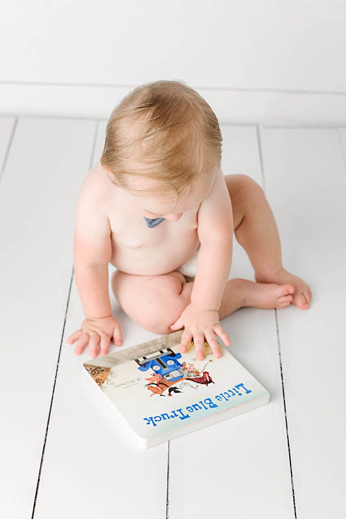 Baby sitting on white floor reading Little Blue Truck board book, viewed from above.