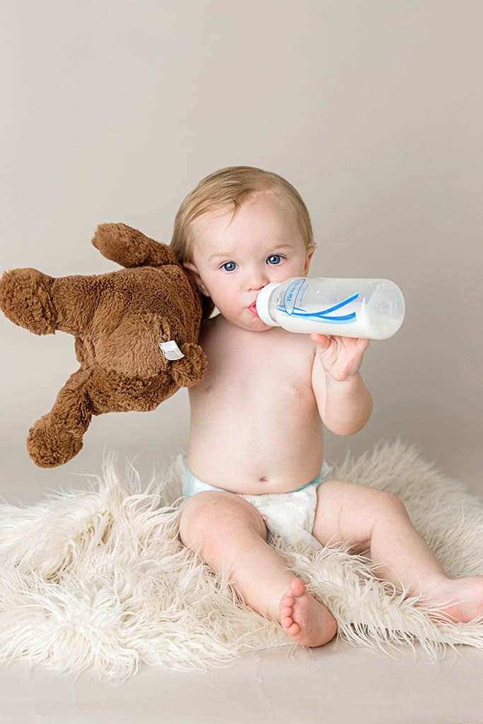Baby in diaper drinking from bottle while holding a brown teddy bear, sitting on fluffy white rug.