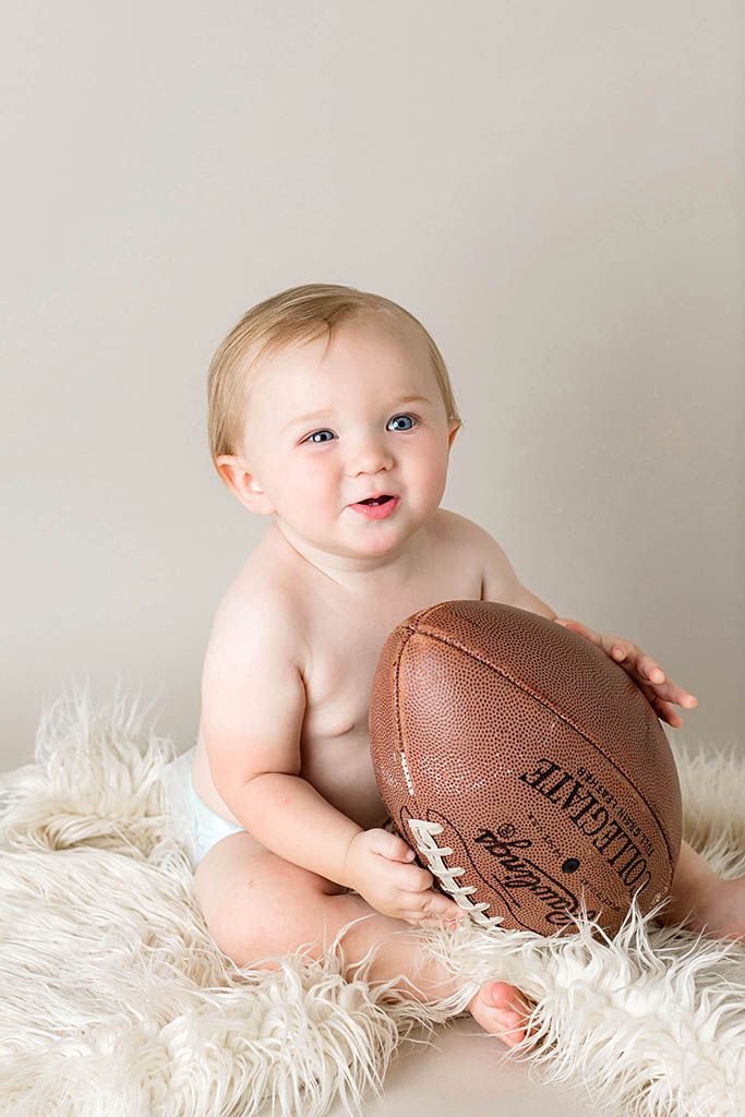 Wildomar baby photographer photo of smiling baby in diaper holding a football while sitting on a fluffy white rug.