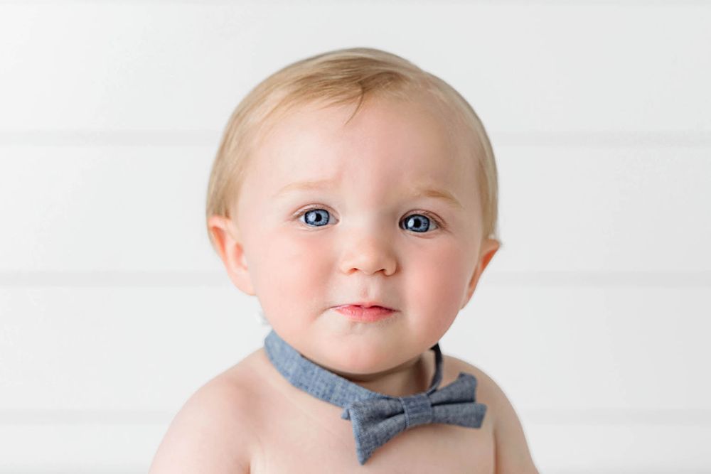 Adorable baby boy with blue eyes wearing a gray bow tie bib against a white background.