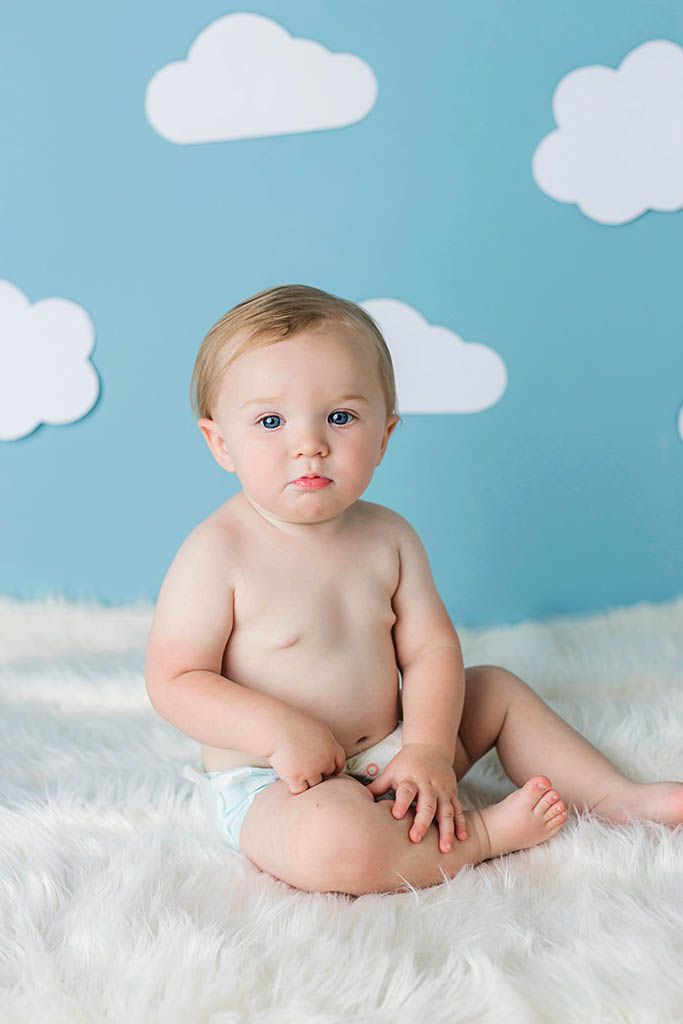 Cute baby boy sitting on white fur rug with blue sky and white clouds backdrop, by Wildomar California baby photographer.